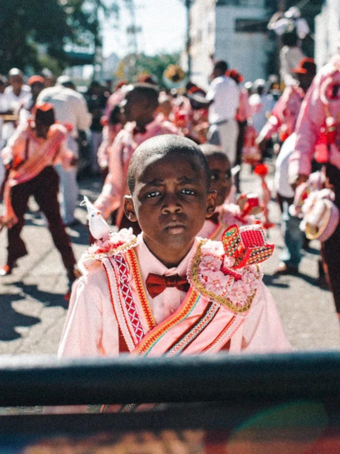 A boy in a pink carnival costume and bow tie, a parade behind him